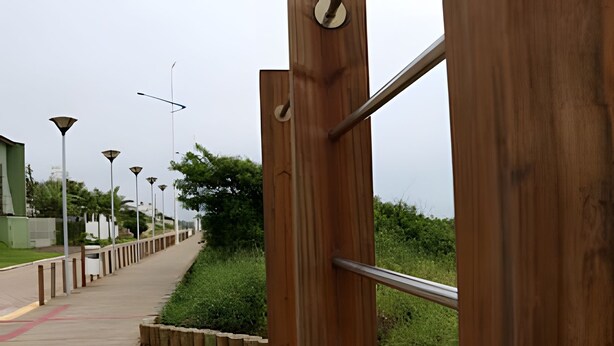 A wooden pedestrian bridge with metal railings, leading to a pathway with street lamps and greenery. - Curta Praia do Quilombo - Palladium (Penha)