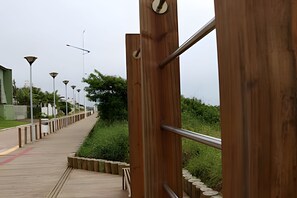 A wooden pedestrian bridge with metal railings, leading to a pathway with street lamps and greenery. - Curta Praia do Quilombo - Palladium  (Penha)