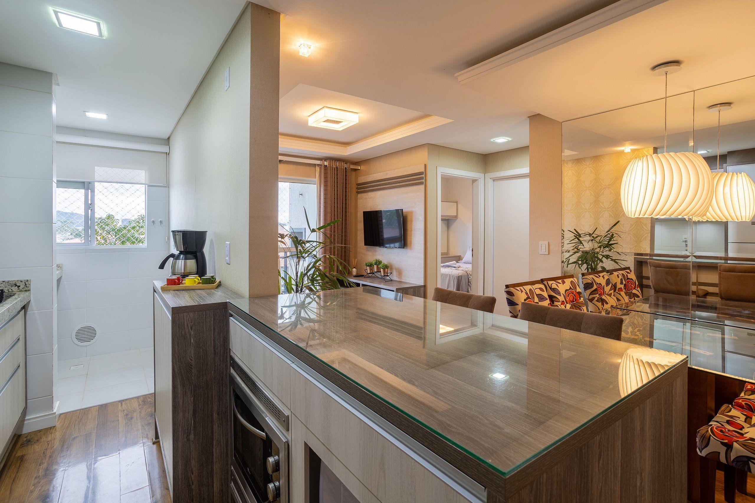 Une cuisine moderne dotée d’un îlot central en verre, un coin repas avec un canapé et des fauteuils, et un salon avec une télévision et un éclairage décoratif.