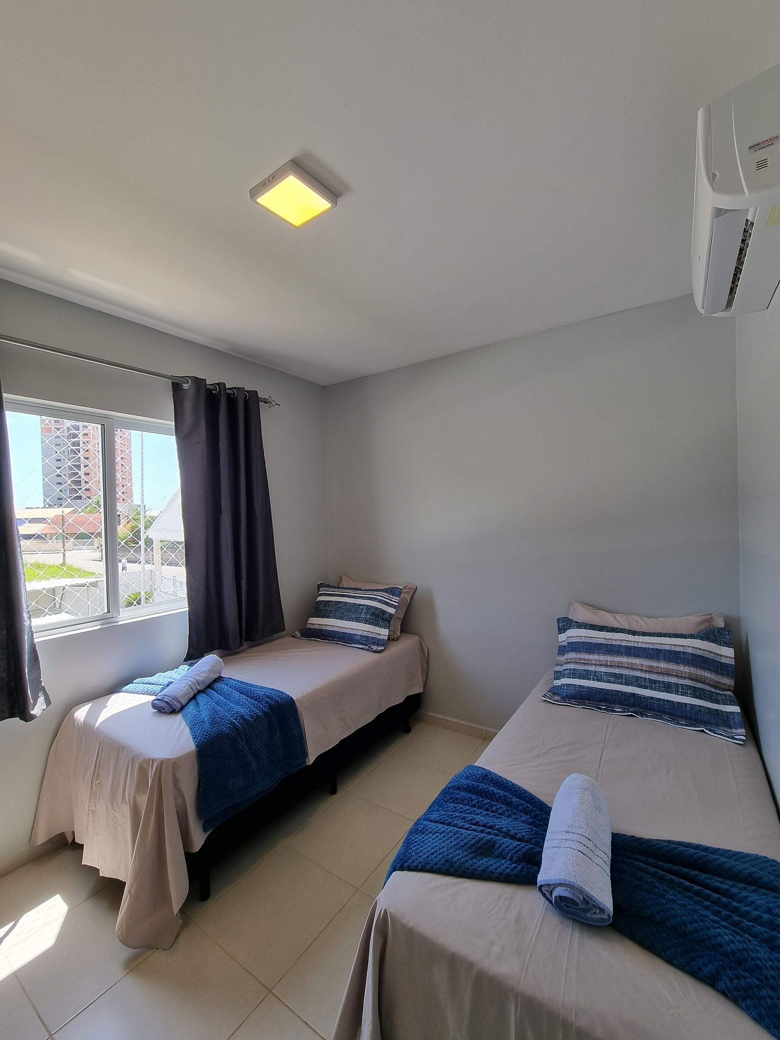 A room with two beds, a window with curtains, a ceiling light, and an air conditioning unit.