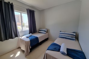 A small, clean room with two beds, a window with curtains, and a view of buildings outside. - Curta Praia do Quilombo - Palladium  (Penha)