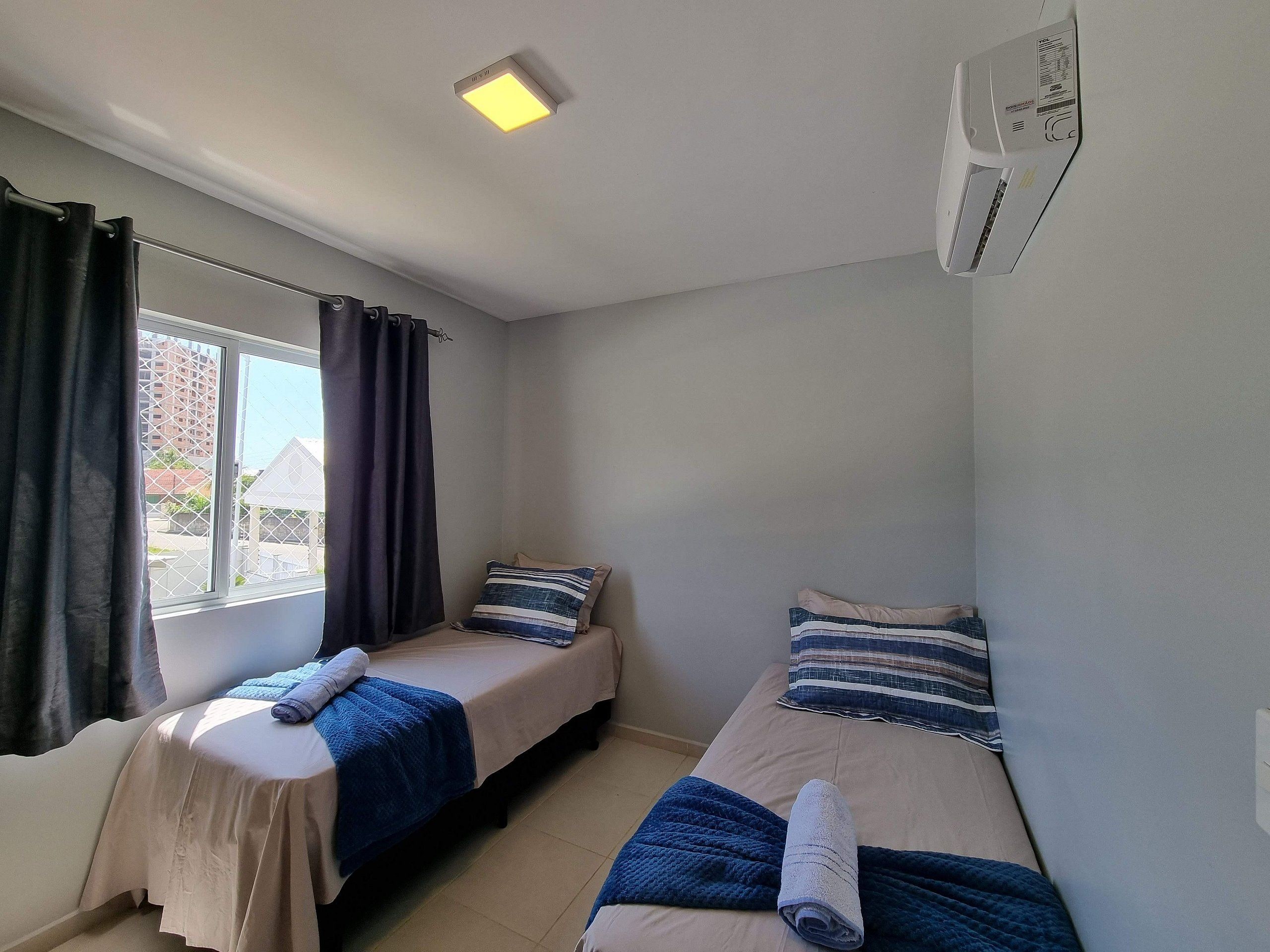 Quarto pequeno e limpo com duas camas, uma janela com cortinas e uma luminária no teto.