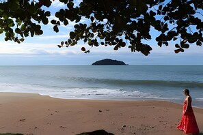 A beach with a person in a red dress, calm sea, and an island in the distance. - Curta Praia do Quilombo - Palladium  (Penha)
