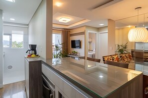 A modern kitchen with a glass island, a dining area with a sofa, and a living room with a TV and plants. - Curta Praia do Quilombo - Palladium  (Penha)