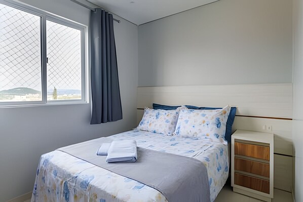 Un dormitorio con cama, ventana con cortinas y mesita de noche.