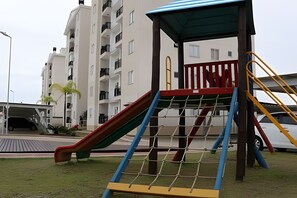A playground with a slide and climbing net, surrounded by a building with balconies and a covered area.
