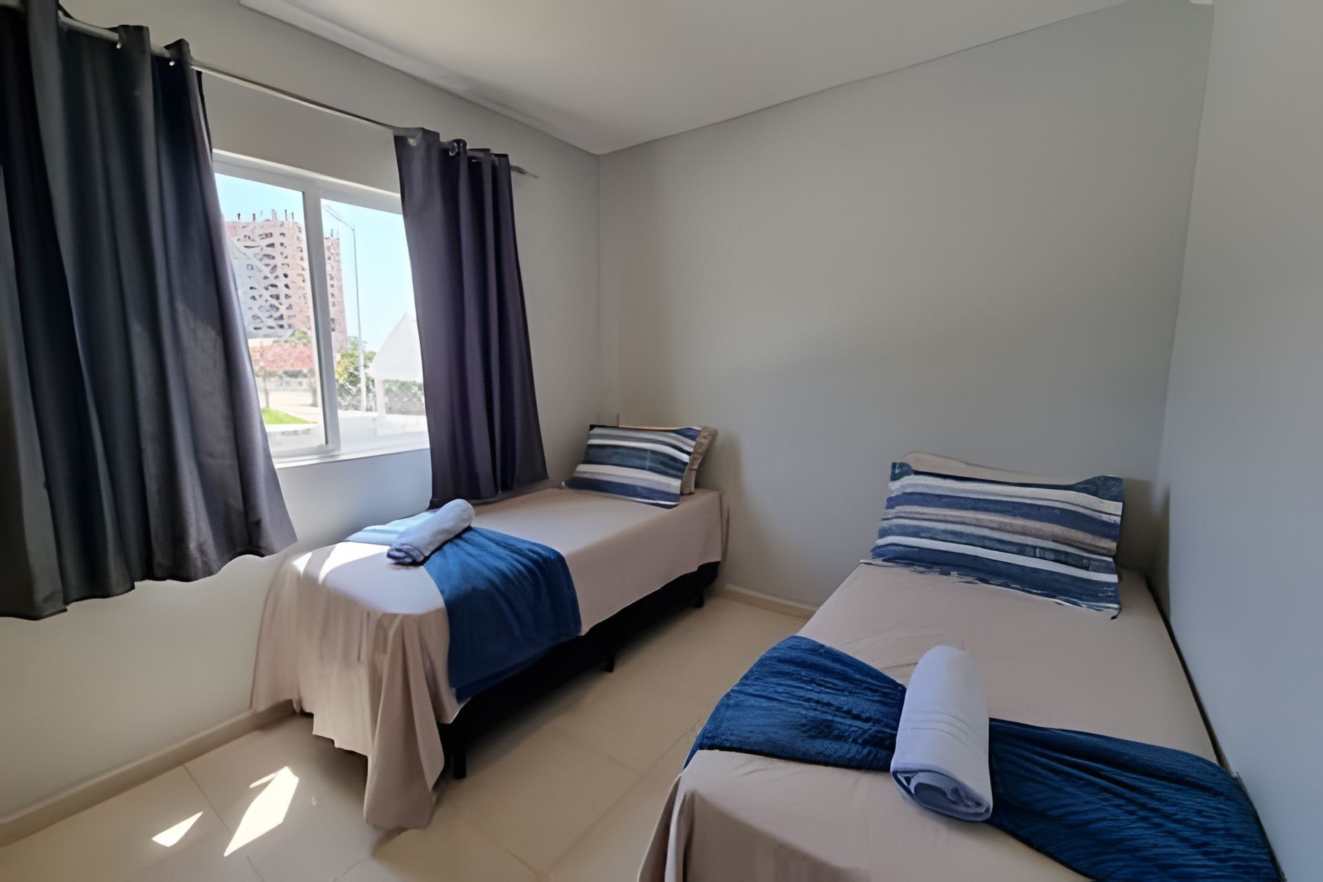 Una habitación pequeña y limpia con dos camas, una ventana con cortinas y vistas a edificios.