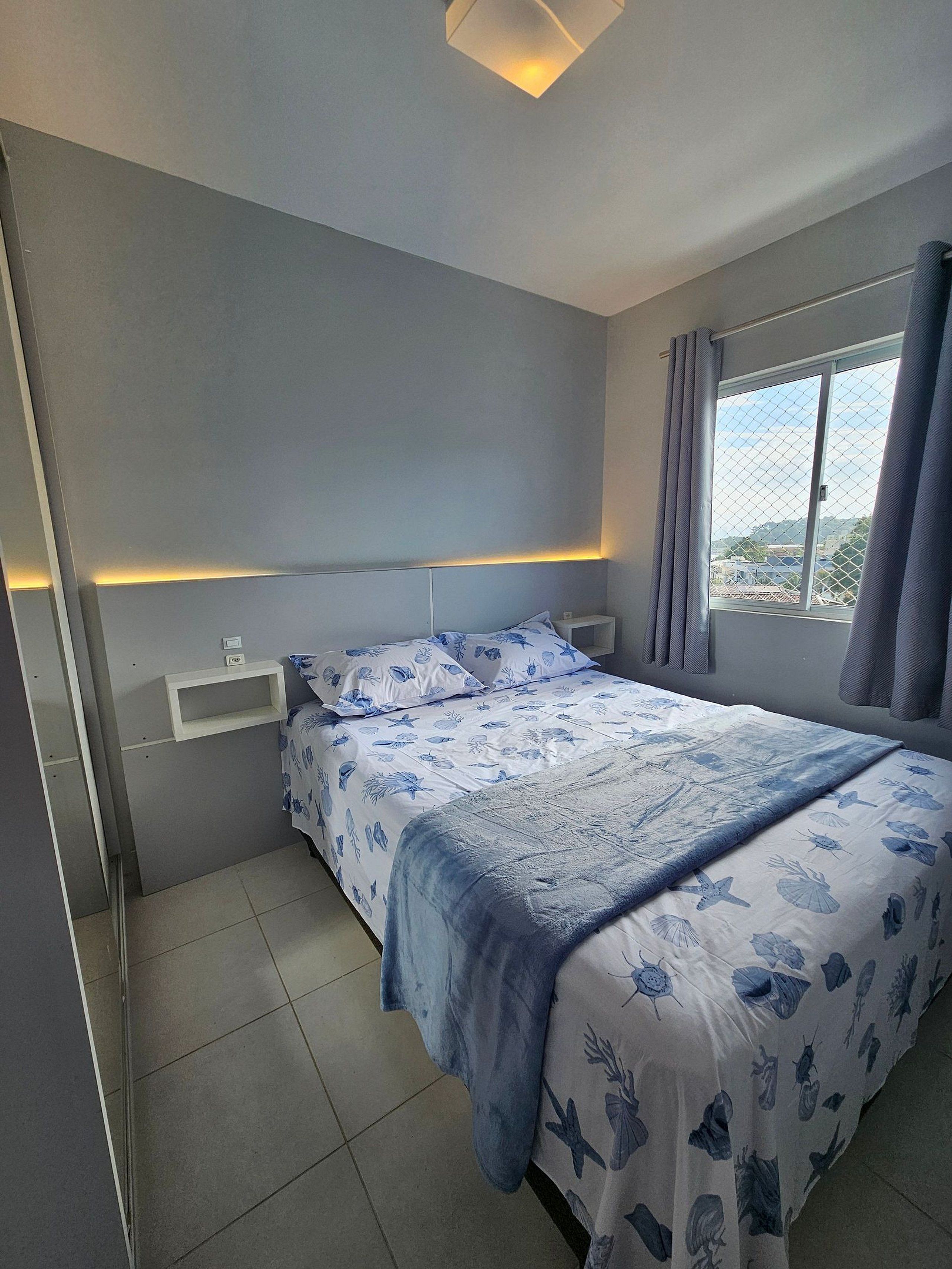 A bedroom with a bed, a bedside table, a window with curtains, and a view of the outside.