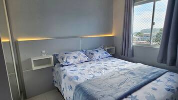 A bedroom with a bed, a bedside table, a window with curtains, and a view of the outside.
