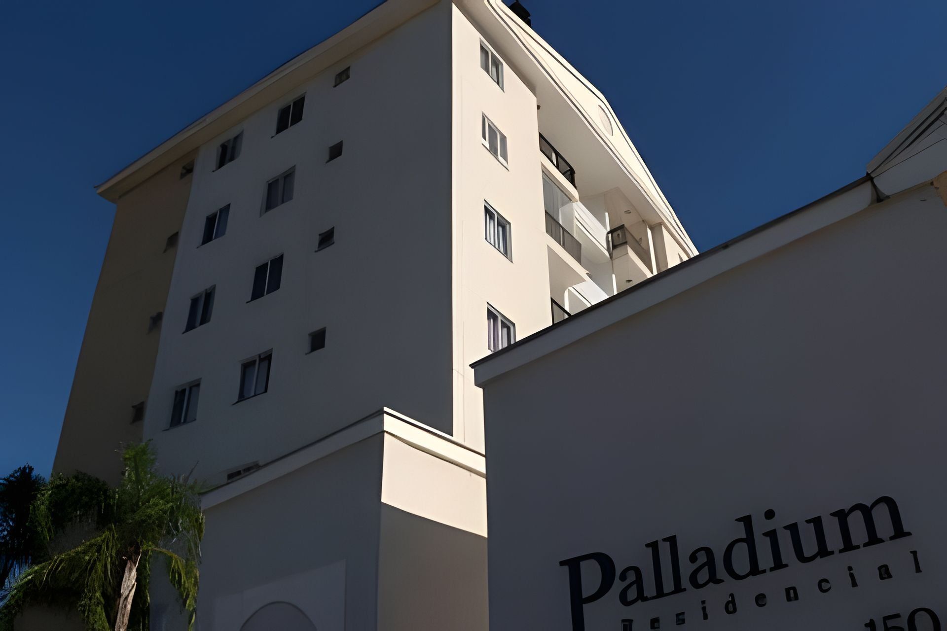 A tall, multi-story building with a white facade and numerous windows. In the foreground, there is a sign with the text "Palladium Residencial".