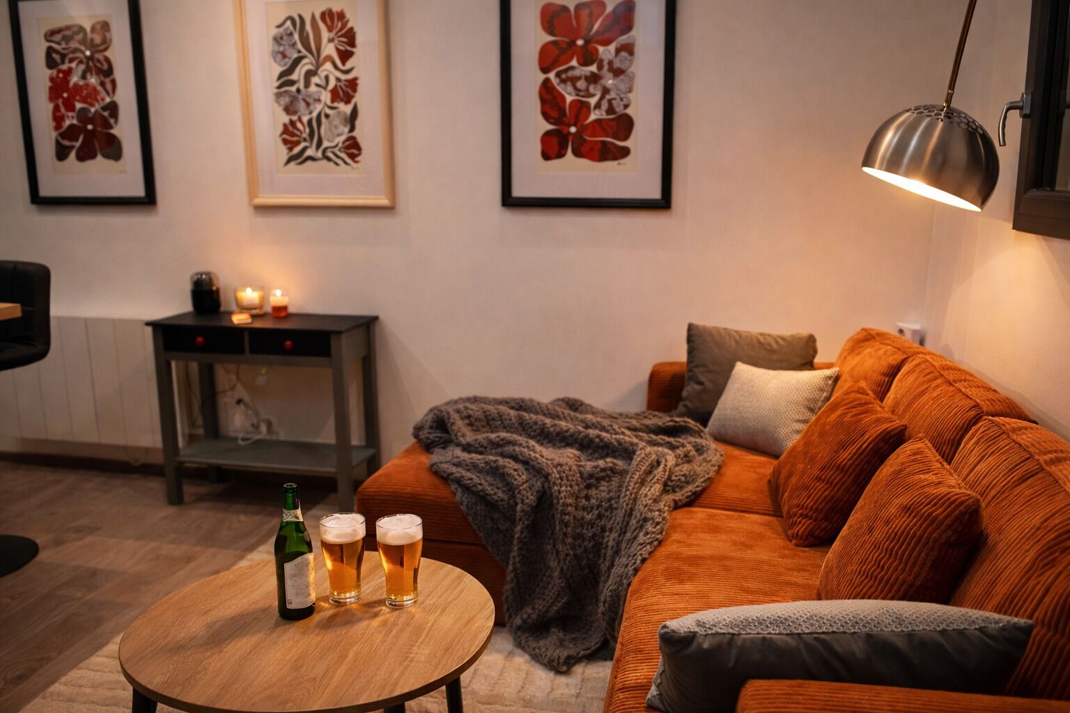 Ein gemütliches Wohnzimmer mit einer Couch, einem Couchtisch mit Getränken, einem Beistelltisch mit Kerze und gerahmten Kunstwerken an der Wand.