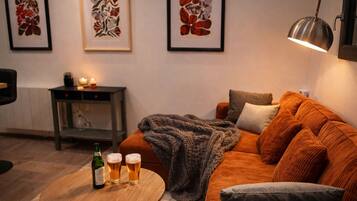 Ein gemütliches Wohnzimmer mit einer Couch, einem Couchtisch mit Getränken, einem Beistelltisch mit Kerze und gerahmten Kunstwerken an der Wand.