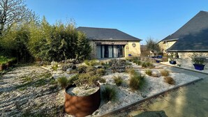 A stone house with a dark roof, surrounded by a gravel garden with various plants and a metal planter.