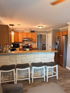 A kitchen with a stone island, wooden cabinets, a stainless steel refrigerator, and a ceiling fan. - Howdy House (College Station)