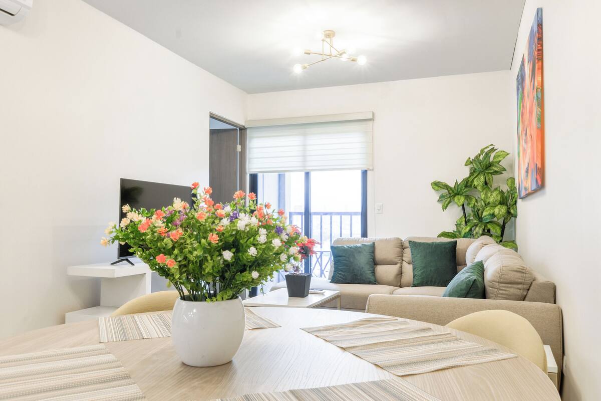 A modern living room with a sofa, a dining table with placemats, a potted plant, and a television.