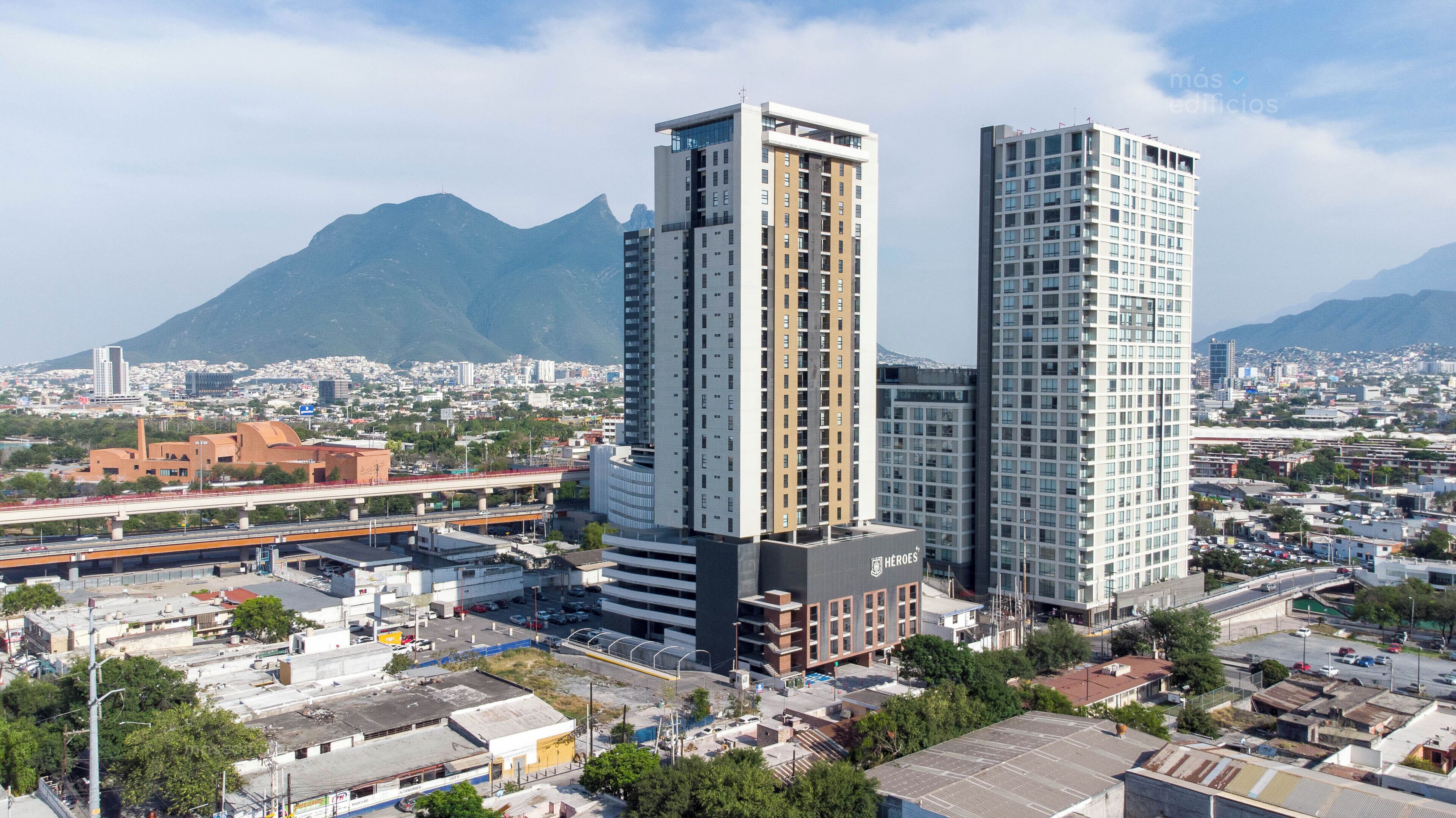 A modern high-rise building with a prominent glass facade, situated in an urban area with a mountain backdrop.