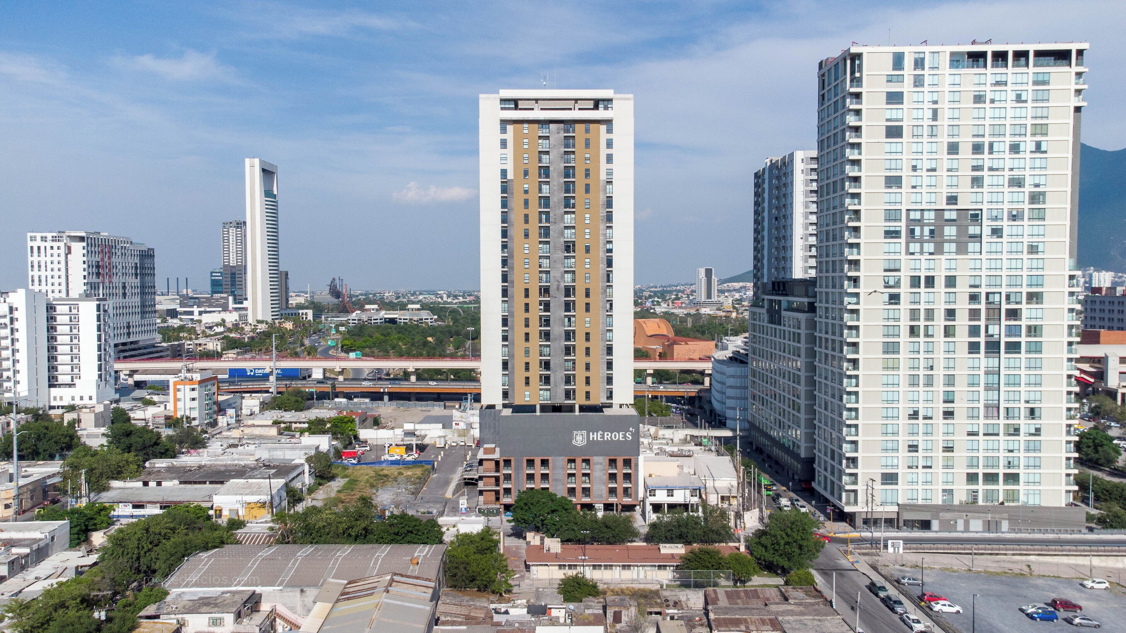 A high-rise building with a central vertical feature, surrounded by other high-rise buildings, with a clear sky and urban landscape in the background.