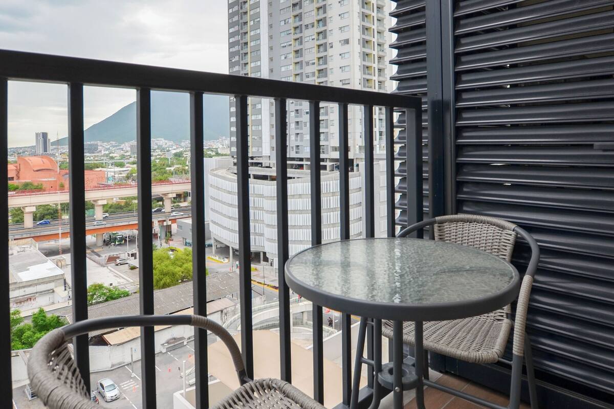 A balcony with wicker chairs and a glass table, overlooking a cityscape with tall buildings and a mountain in the distance.