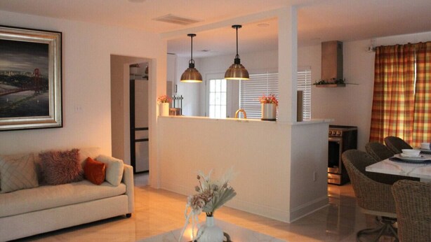 A modern living room with a sofa, a dining area with wicker chairs, a kitchen with a refrigerator, and pendant lights.