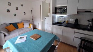 Een knus woongedeelte met een kitchenette, een bank met kussens, een eettafel met een blauwe tafelkleed en een klein houten snijplank.