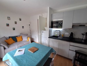 A compact living space with a kitchenette, a sofa with cushions, a dining table with a blue tablecloth, and a small wooden cutting board. - Appart' Pyrénées Nature, comfortable, with terrace and small garden (Esquièze-Sère)