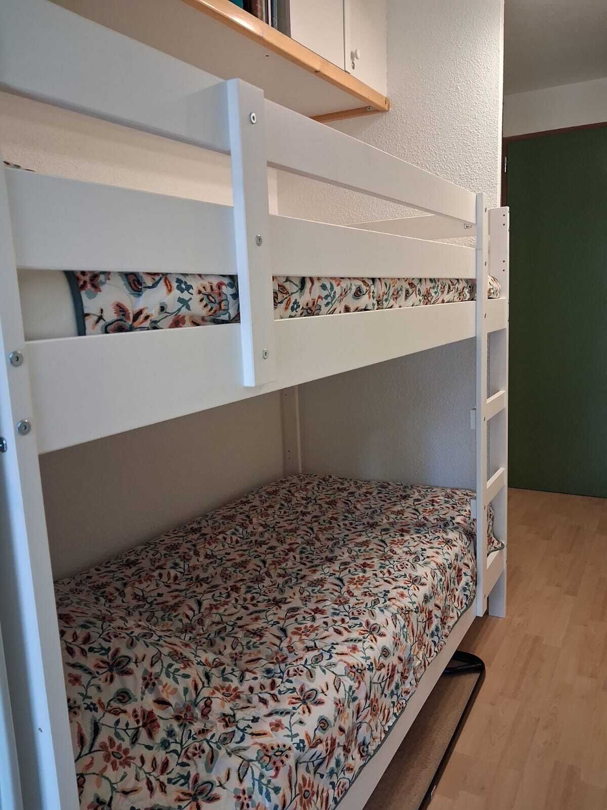 Uma cama beliche com roupa de cama florida, uma prateleira de madeira acima e uma parede verde.