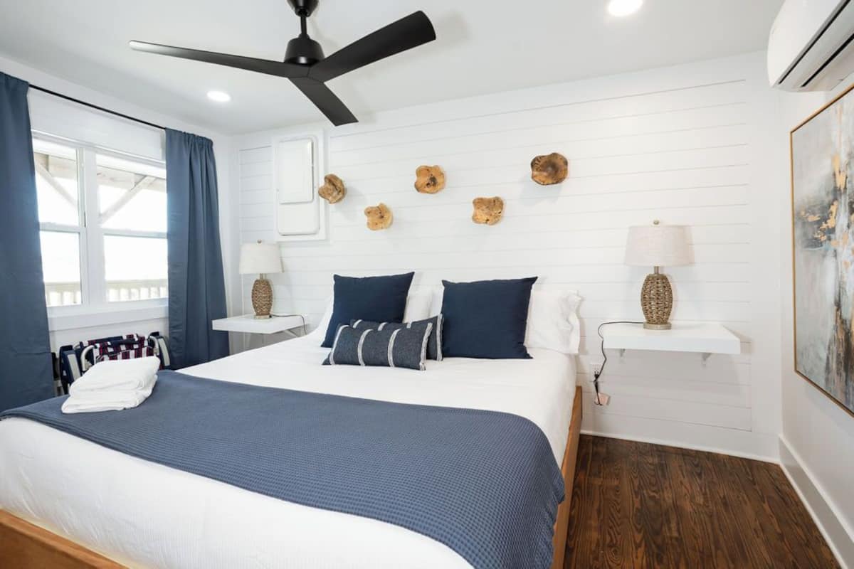 Un dormitorio con cama, ventilador de techo, ventana con cortinas, decoración en la pared y un cuadro.