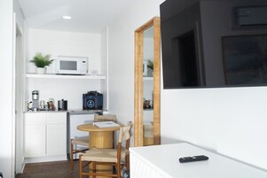 A compact room with a kitchenette, a round table with two chairs, a large mirror, and a remote control on a white surface.