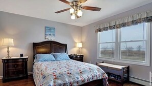 A bedroom with a wooden bed, a ceiling fan, a nightstand with a lamp, a window with a view, and a bench by the window.