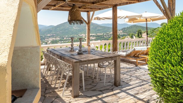 Un pati exterior amb taula i caires de fusta, un teulat de palla i vistes al camp.