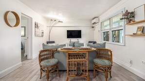 Een ruimtelijk ingerichte zithoek met een eethoek, bestaande uit rieten stoelen, een ronde tafel, een grijze bank en een wandgemonteerde flatscreen televisie.
