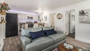 Een moderne woonkamer met een grijze leren bank, een keuken op de achtergrond en een deur naar een andere kamer.