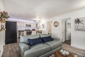 A modern living room with a grey leather sofa, a kitchen in the background, and a door leading to another room. - Vacation Vibes Escape (Cape Canaveral)