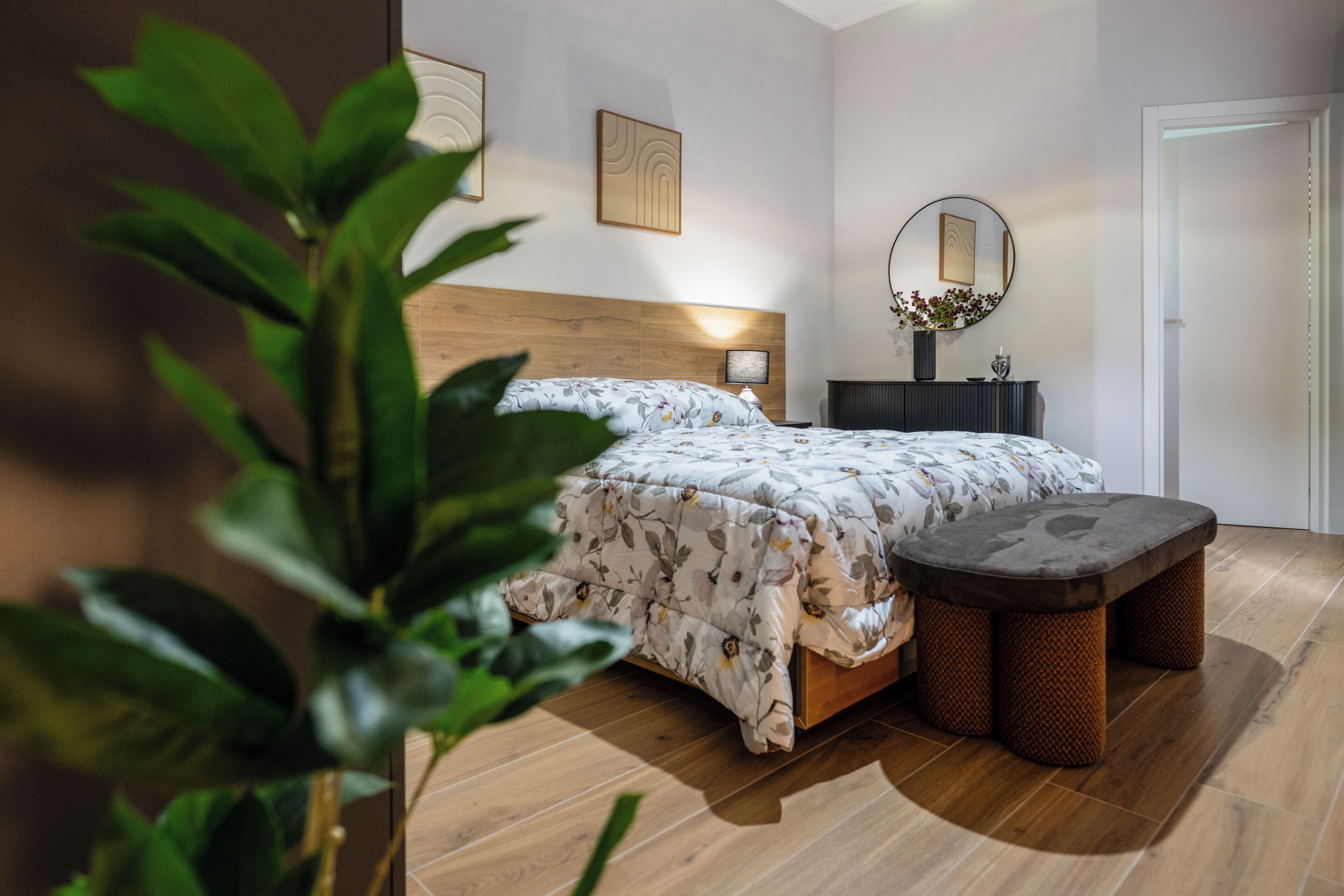 A bedroom with a wooden headboard, a bed with floral bedding, a bedside table with a lamp, a round mirror, and a small bench.