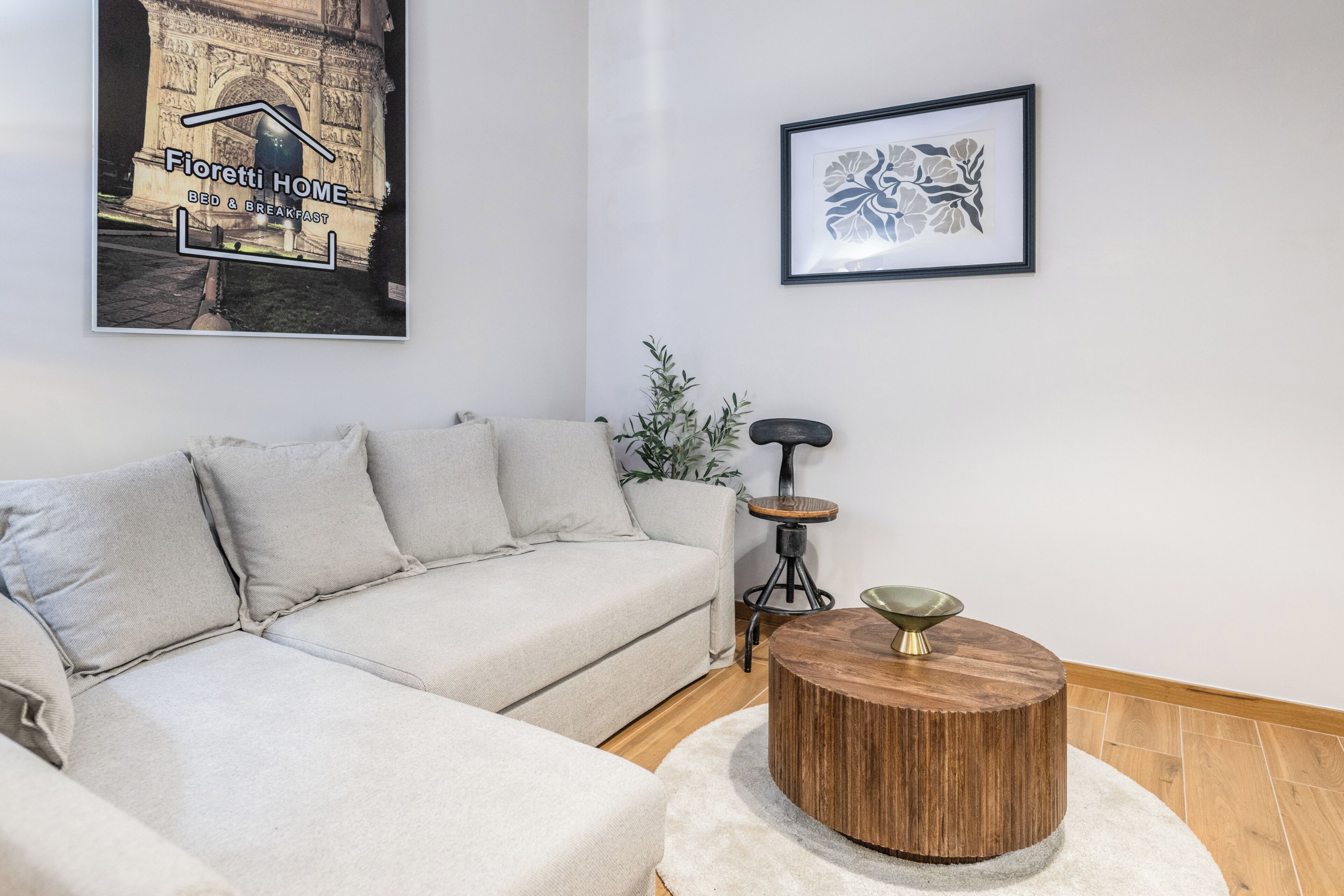 Una sala de estar con un sofá, una mesa de centro redonda de madera, una obra de arte floral enmarco y un cartel en la pared.