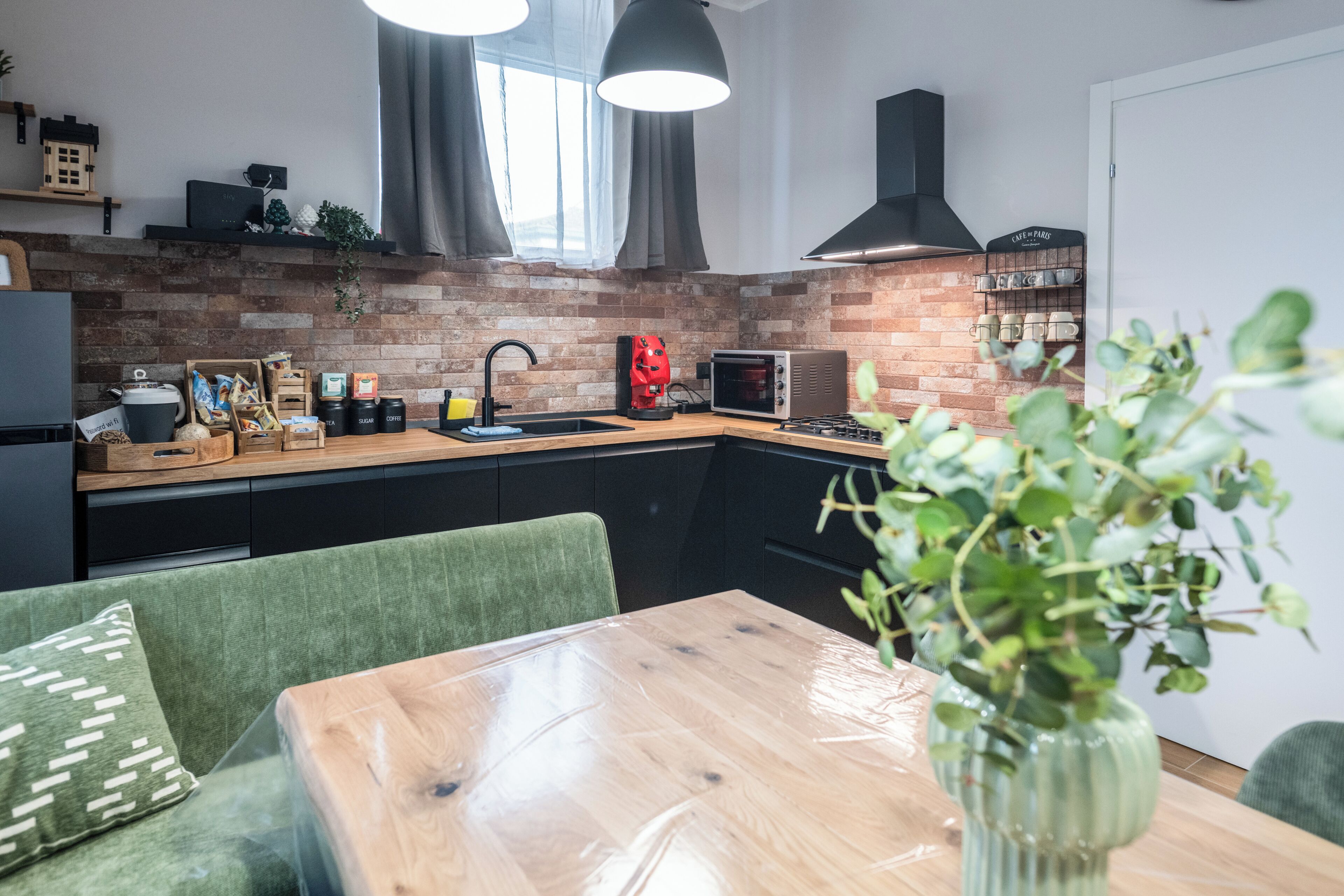 Une cuisine moderne avec un mur recouvert de briques, des meubles de couleur noire, une table à manger en bois et un canapé vert.