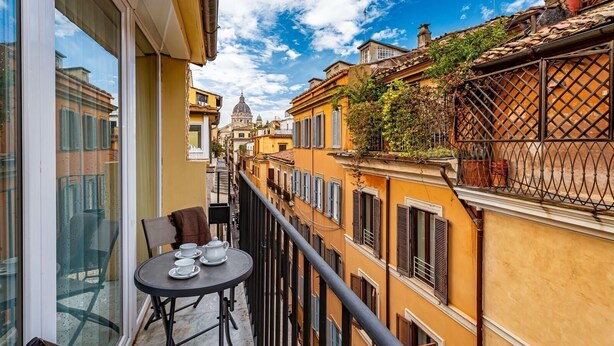 Un balcón con una mesa lista para dos personas, con vista a una calle estrecha flanqueada por edificios amarillos y una estructura abovedada al fondo.
