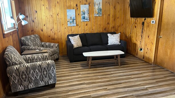 Une pièce aux murs recouverts de panneaux de bois, avec un canapé noir, des fauteuils à motifs, une table basse blanche et un téléviseur à écran plat fixé au mur.