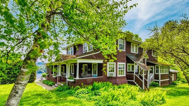 A two-story house with a porch, surrounded by lush green grass and trees. - Walk to Main Street & Mirror Lake (Lake Placid)