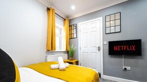 Een hotelkamer met een bed, een aan de muur bevestigde televisie, een raam met gele gordijnen, een witte deur en een klein tafeltje met een plant.