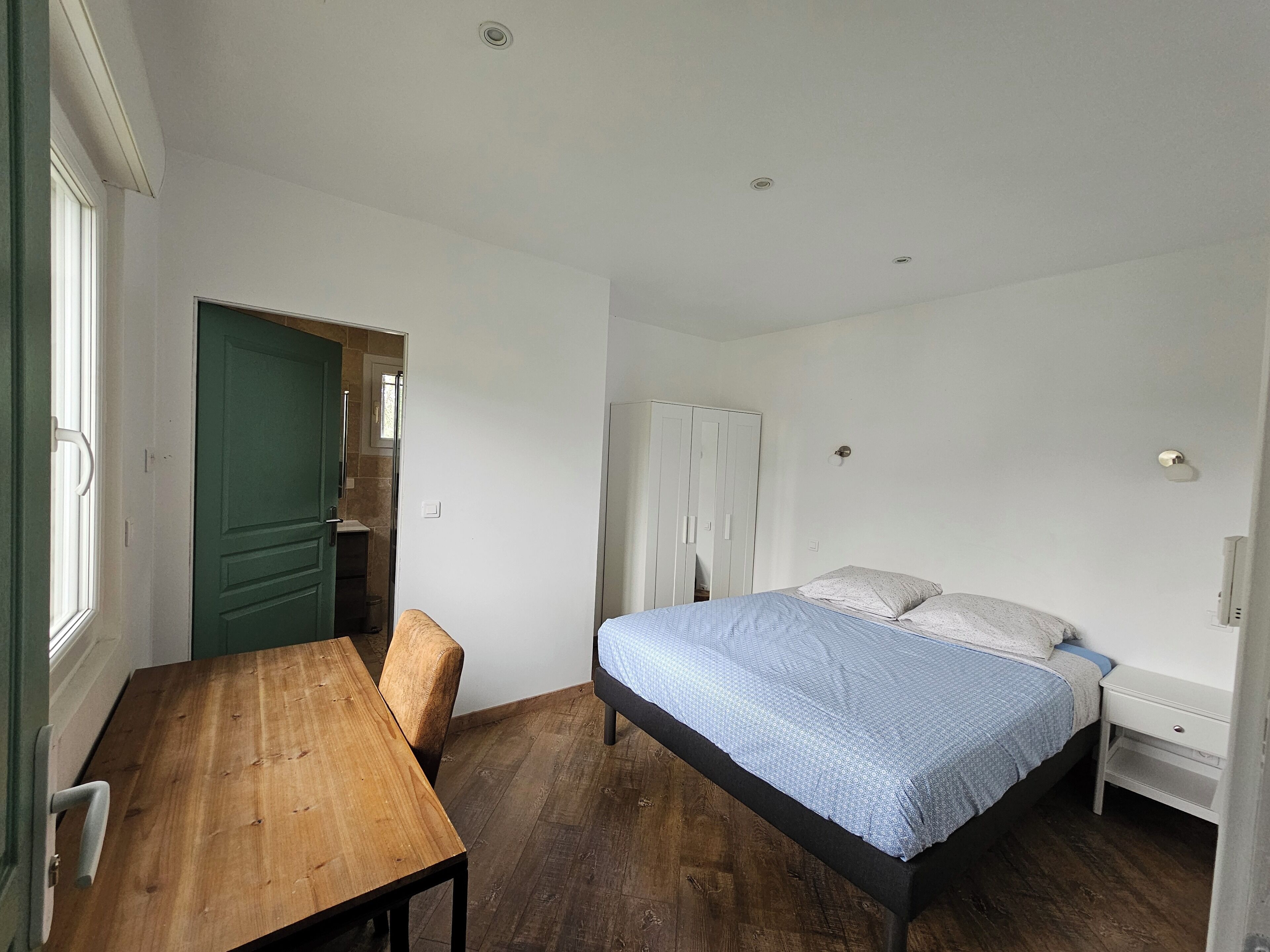 Une chambre avec une table en bois, un lit recouvert d’une couverture bleue et une porte verte.