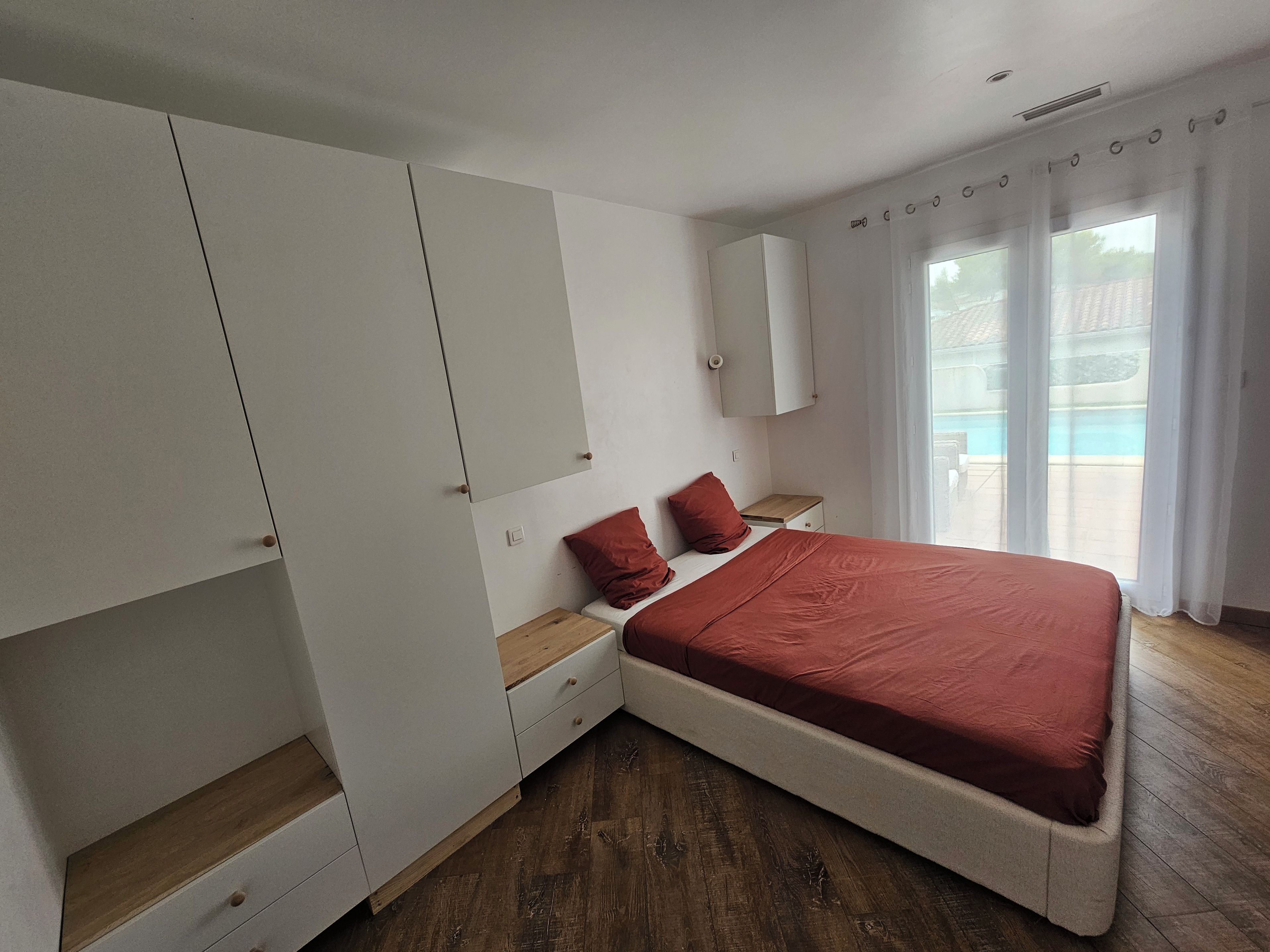 A bedroom with a bed, a wardrobe, a bedside table, and a window with curtains.