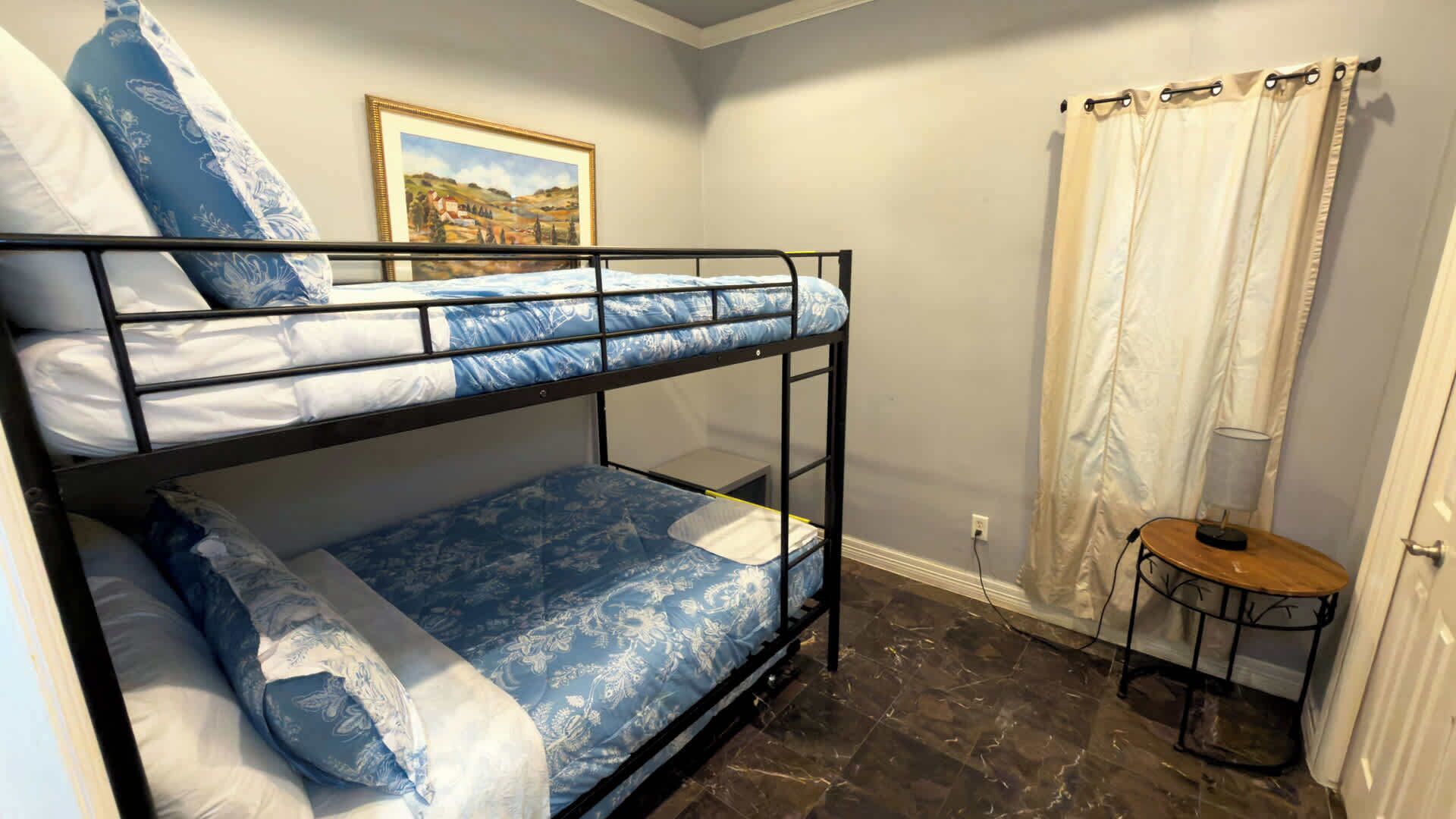 A bunk bed room with a framed picture on the wall, a small table with a lamp, and a curtain on the window.
