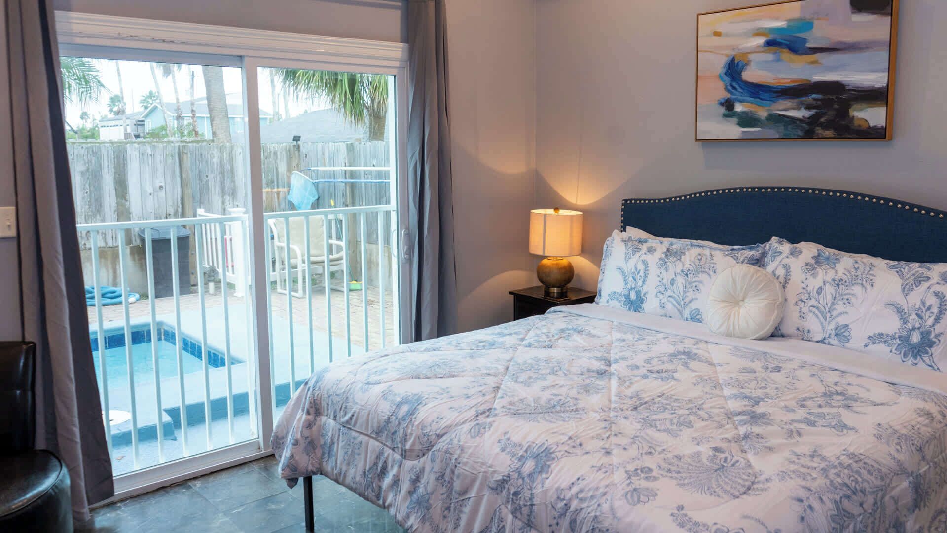 Un dormitorio con cama, mesita de noche, lámpara y un cuadro en la pared.