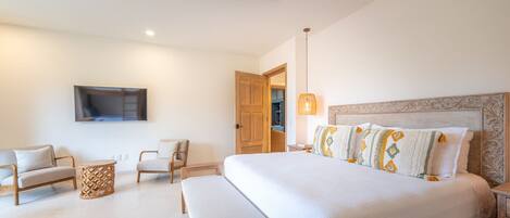 Un dormitorio con una cama grande, dos sillones, una mesita pequeña, un ventilador de techo, un televisor montado en la pared y una puerta que da a otra habitación.
