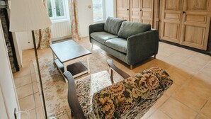 A living room with a sofa, a coffee table, a floor lamp, and a patterned ottoman. There is a window with curtains, a radiator, and wooden cabinets in the background. - Bright English-style cottage apartment (Mazet-Saint-Voy)
