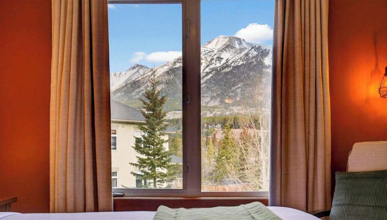 Une chambre d’hôtel avec un lit, une fenêtre avec des rideaux et une vue sur des montagnes enneigées.