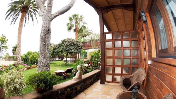 Un porche de madera con suelo de piedra, mobiliario de mimbre y vistas a un jardín bien cuidado con palmeras y césped.