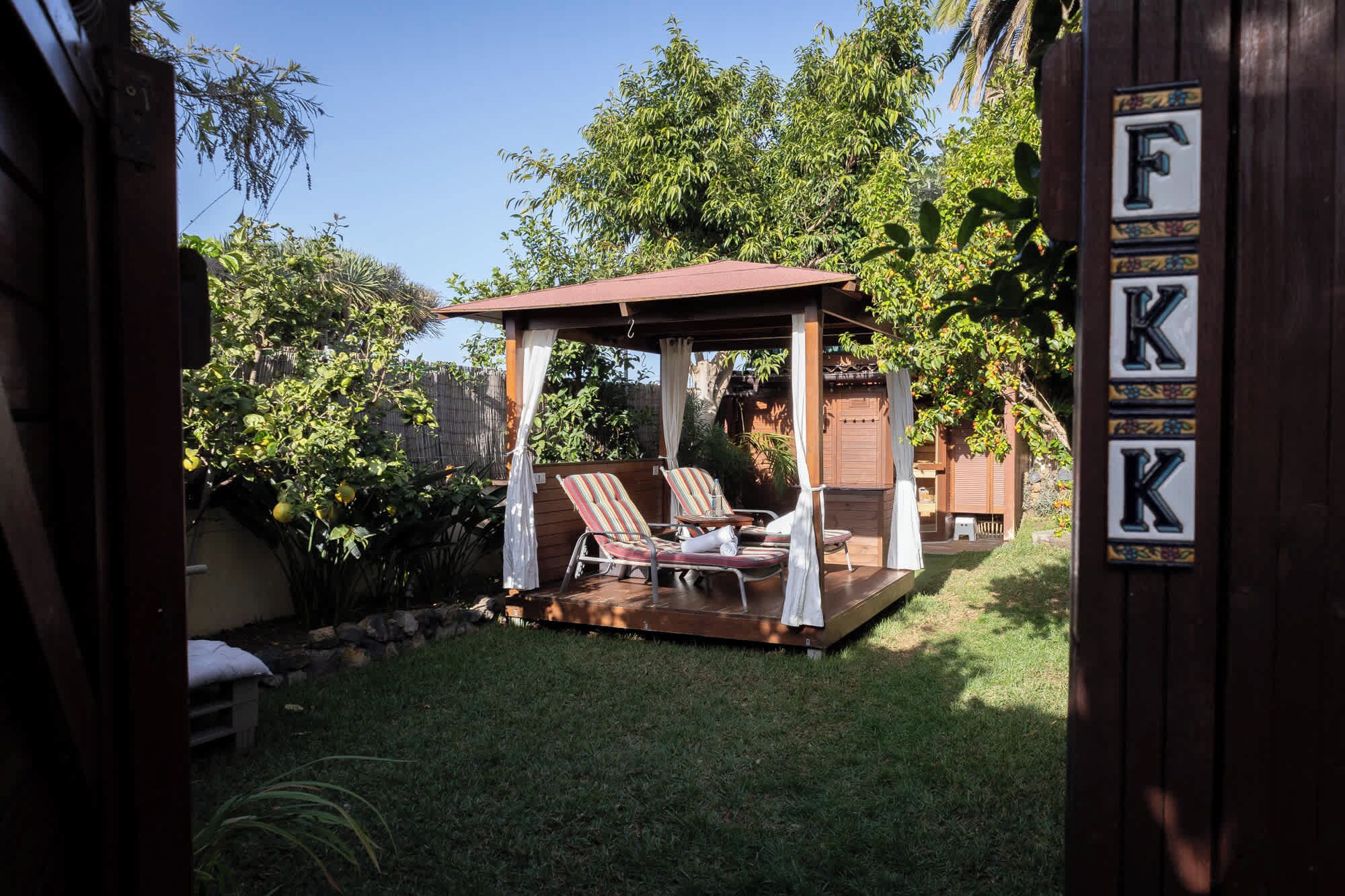 Ein überdachter Patio-Bereich mit zwei Liegestühlen und einem kleinen Tisch, umgeben von üppiger Bepflanzung und einem Holzzaun.