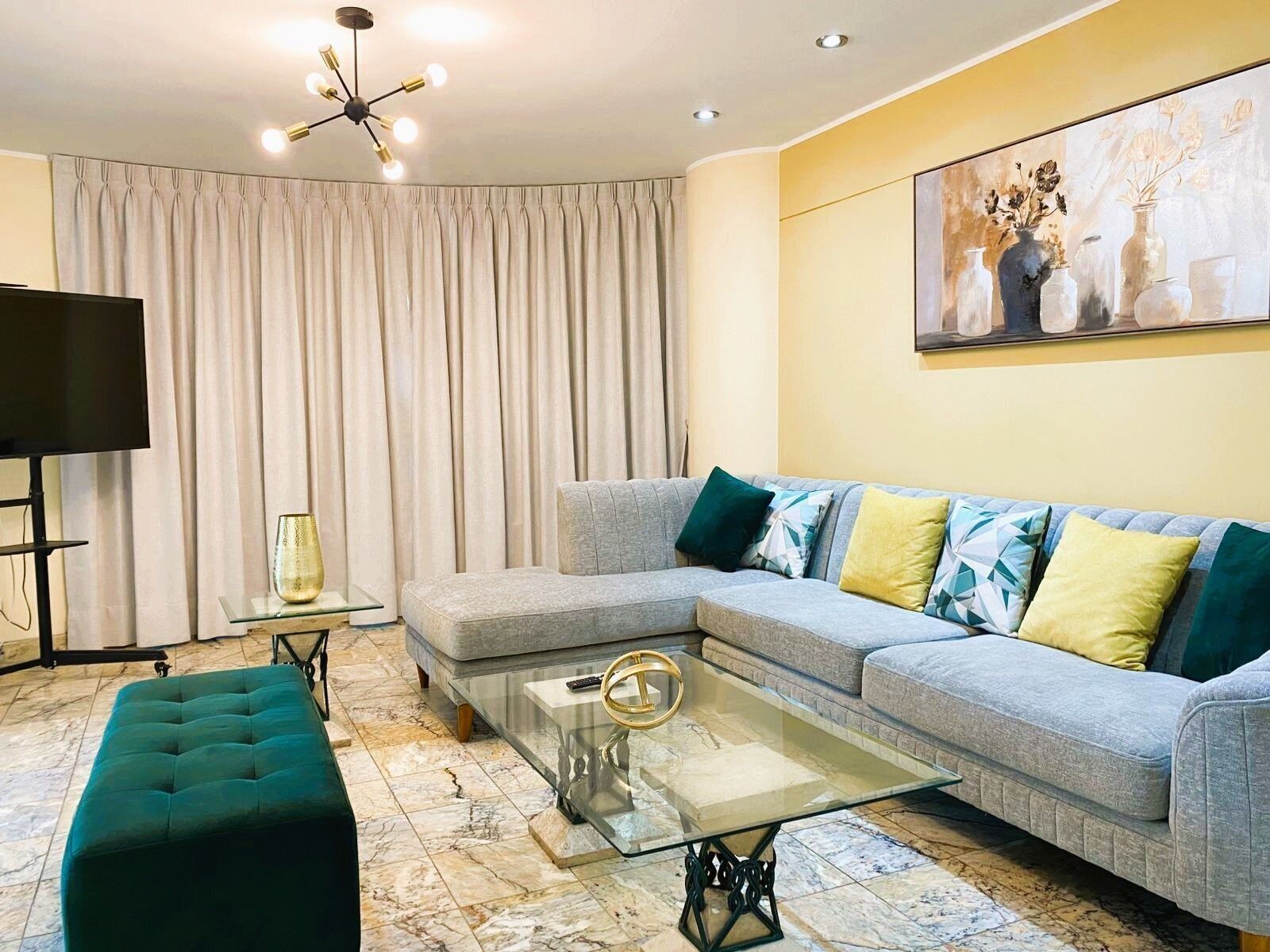 Una sala de estar con un sofá modular, una mesa de centro de vidrio, un reposapiés color turquesa, un televisor de pantalla plana y un cuadro en la pared.